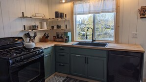 A kitchen with a stove, oven, sink, and cabinets. There are shelves above the counter with various items, and a window with a view of trees. - Magical Northern Michigan Cottage with Hot Tub (Bear Lake)