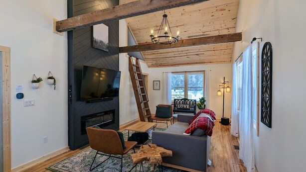 A modern living room with a fireplace, wooden beams, a sofa, and a television. - Magical Northern Michigan Cottage with Hot Tub (Bear Lake)