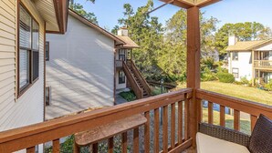 Um deck de madeira com uma cadeira e uma pequena mesa, com vista para uma área residencial com casas e árvores.