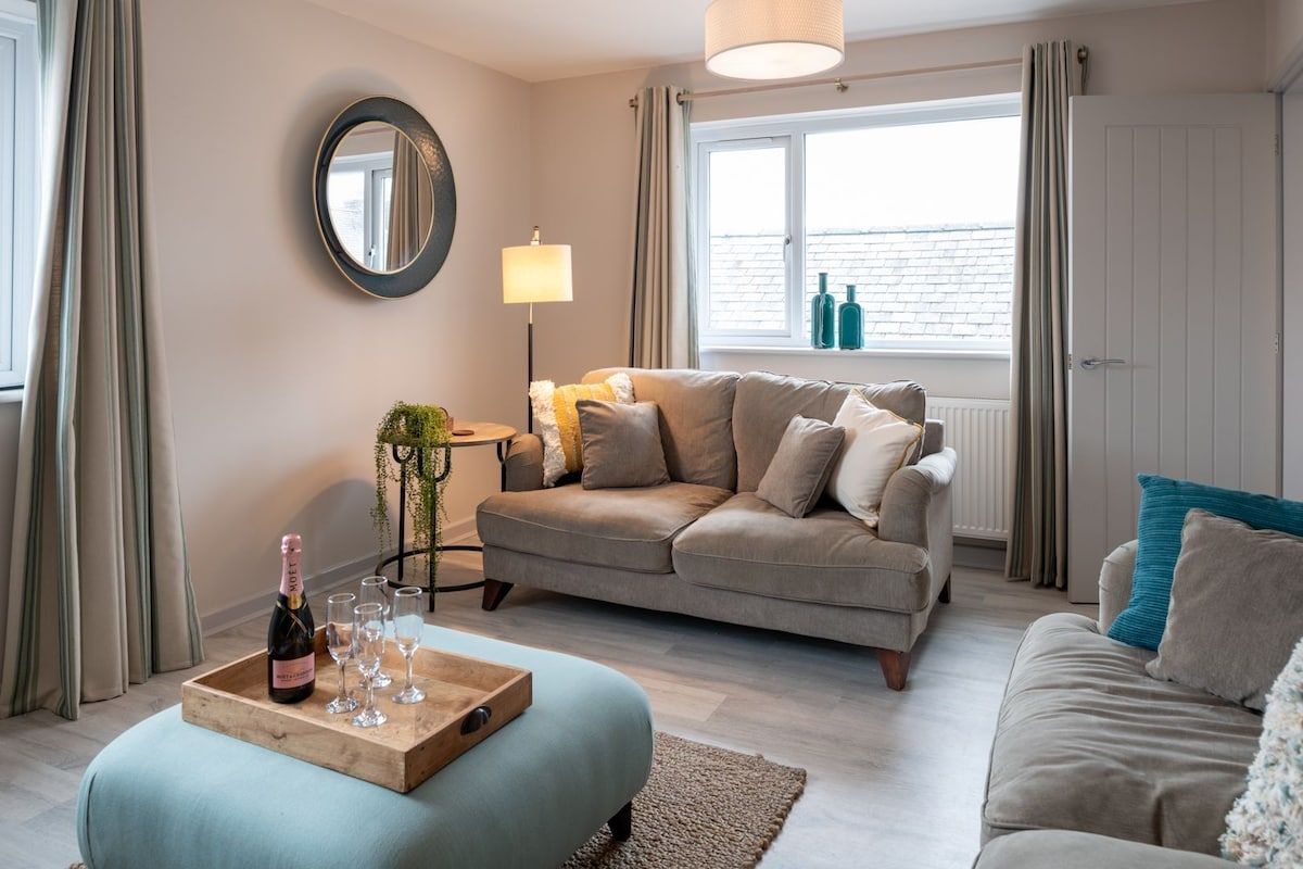 A cozy living room with a sofa, a coffee table with a bottle and glasses, a lamp, and a window with curtains.
