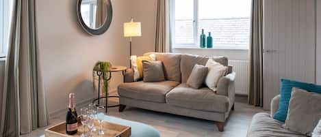 Ein gemütliches Wohnzimmer mit einer Couch, einem Couchtisch mit einer Flasche und Gläsern, einer Lampe und einem Fenster mit Vorhängen.