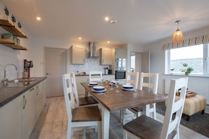 A modern kitchen with white cabinets, a wooden dining table, and a view of the sea.