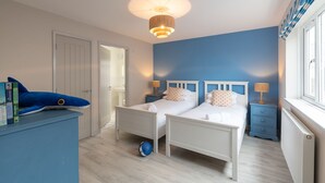 A bedroom with two beds, a blue wall, a ceiling light, a window, and a blue dresser. - Kittiwake Cottage : Stylish 2‑Bed Pet-Friendly Cottage in Rhosneigr | Managed by Anglesey Holiday Le (Rhosneigr)