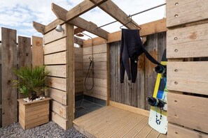 A wooden outdoor shower area with a wooden bench, a potted plant, and a snowboard leaning against the wall.