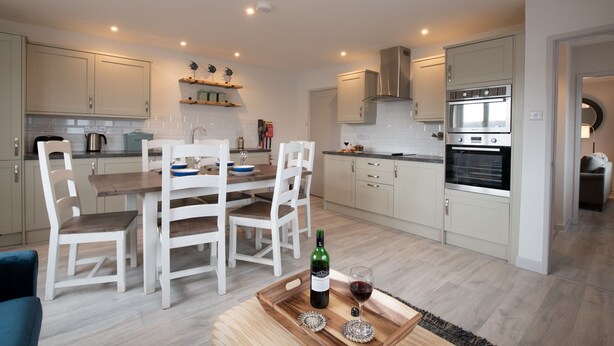A modern kitchen with white cabinetry, a dining table with chairs, and a built-in oven. - Kittiwake Cottage : Stylish 2‑Bed Pet-Friendly Cottage in Rhosneigr | Managed by Anglesey Holiday Le (Rhosneigr)