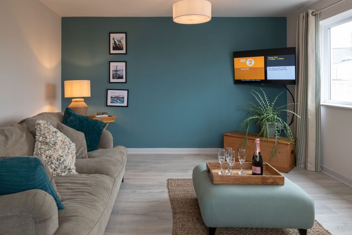 A modern living room with a sofa, a coffee table with glasses and a bottle, a television mounted on the wall, and a window with curtains.