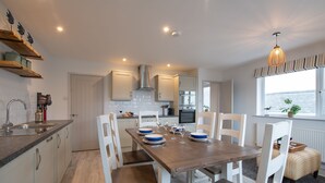 A modern kitchen with white cabinets, a wooden dining table, and a view of the sea through a window. - Kittiwake Cottage : Stylish 2‑Bed Pet-Friendly Cottage in Rhosneigr | Managed by Anglesey Holiday Le (Rhosneigr)
