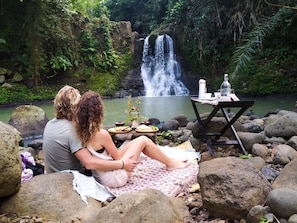 Two people sitting on a blanket by a waterfall, with a table set for a meal in the foreground. - Galalima Glamping (Selemadeg)