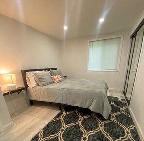 Quarto com cama, mesa de cabeceira, luminária, janela e tapete estampado.