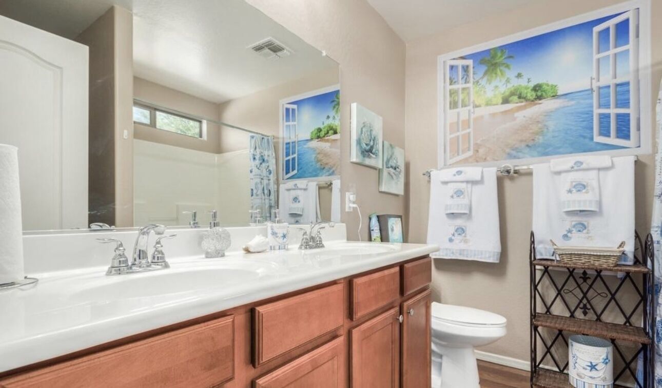 A bathroom with a large mirror, two sinks, wooden cabinets, a toilet, and a shelf with towels.