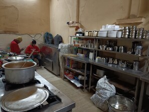 A kitchen with stainless steel appliances, pots, and utensils. There are two people working, one standing and one sitting. - Dalton Hotels  (New Delhi)