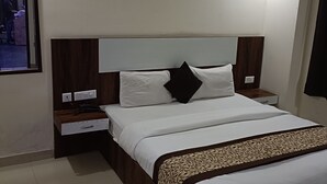 A neatly made bed with white linens, a wooden headboard, and a decorative runner. There is a nightstand with a drawer and a lamp. A ceiling fan is visible above the bed. - Dalton Hotels  (New Delhi)