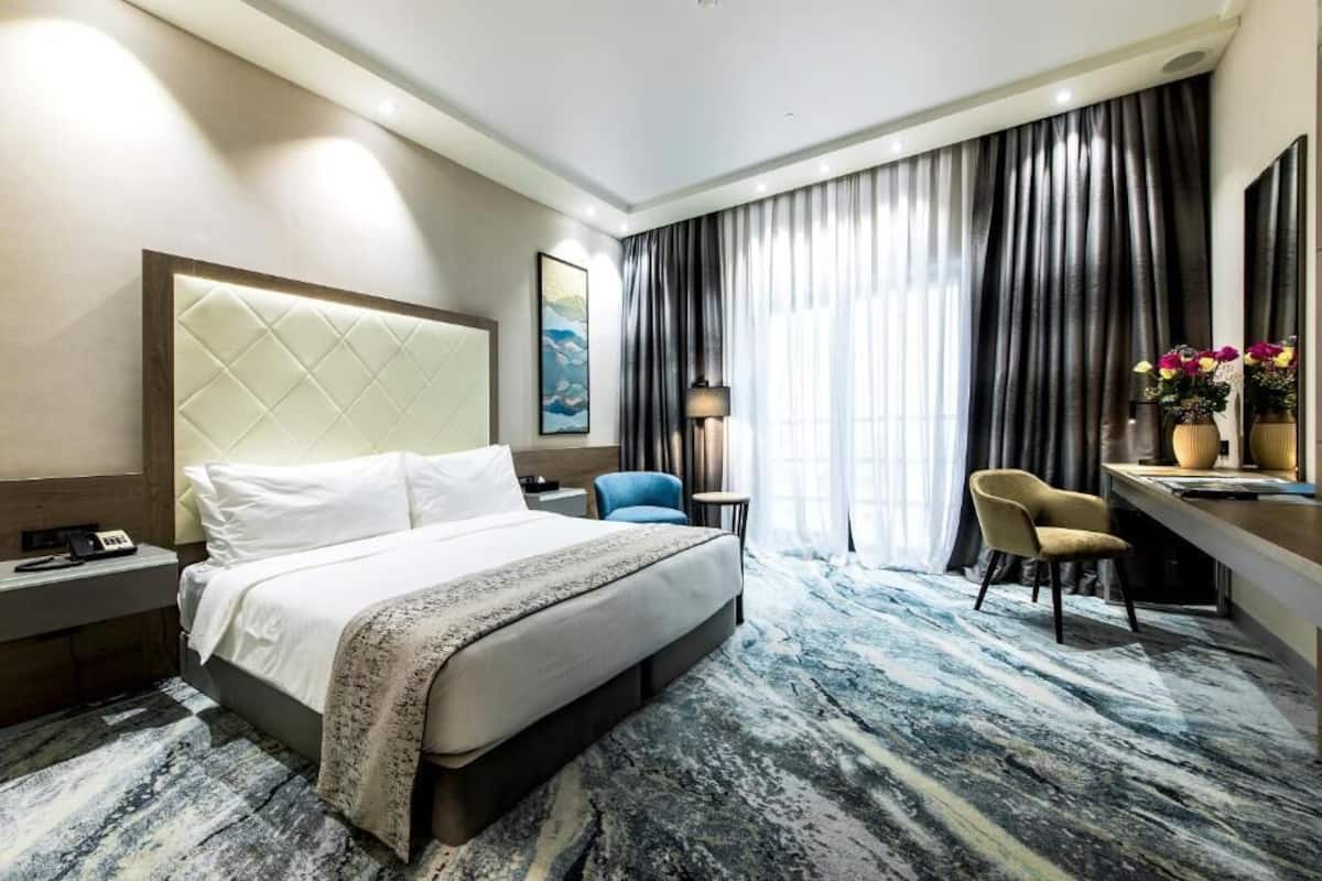 A hotel room with a large bed, a desk with a chair, a nightstand, and a large window with curtains.