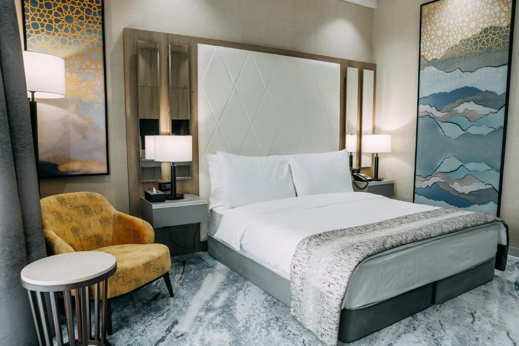 Une chambre d’hôtel avec un grand lit, une petite table ronde, un fauteuil jaune et deux lampes de chevet.