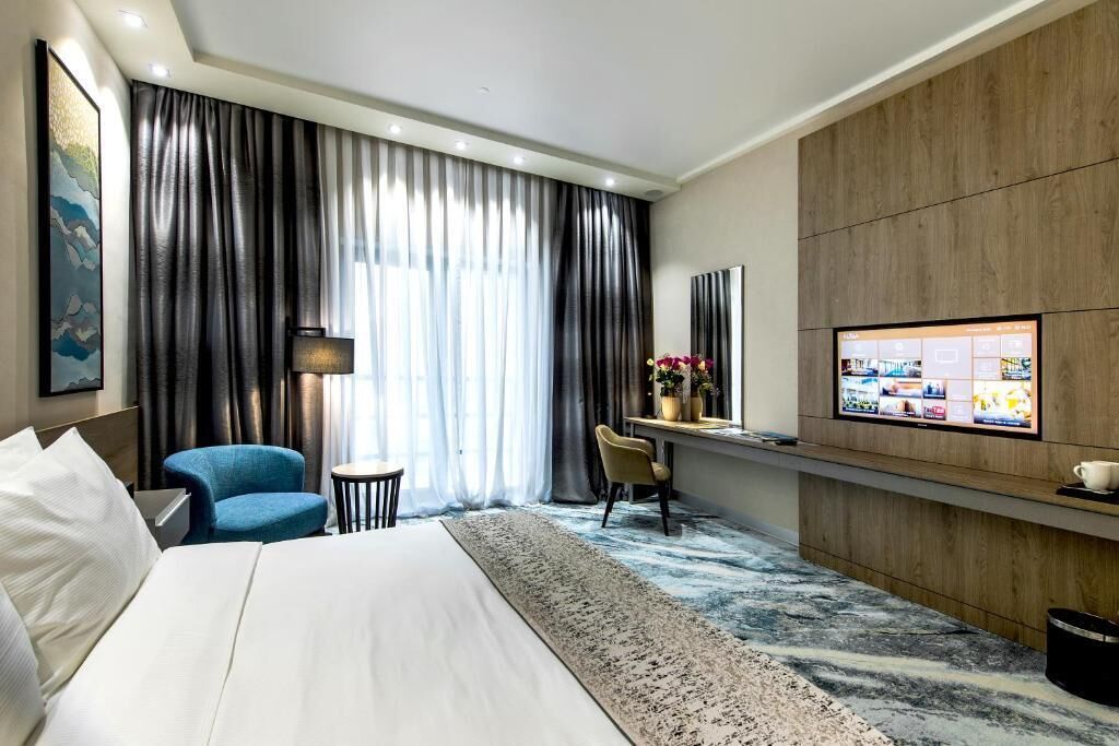 A hotel room with a large bed, a desk with a computer, a chair, a small table, a lamp, and a TV on the wall.