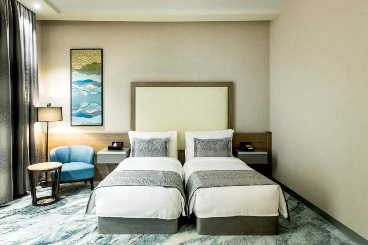 A hotel room with two beds, a desk, a chair, and a lamp.