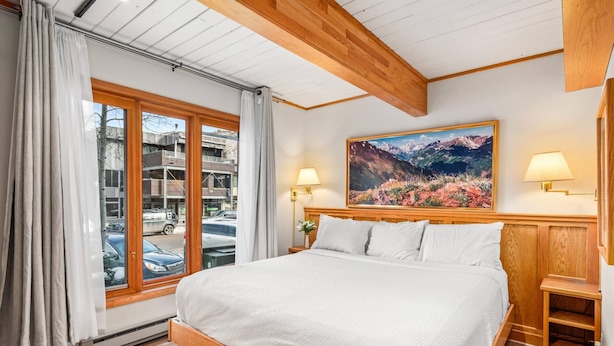 Een slaapkamer met een groot bed, een houten hoofdbord, twee bedlampen, een schilderij en uitzicht op de straat door een raam.
