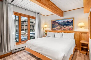 A bedroom with a large bed, wooden headboard, two bedside lamps, a painting, and a view of the street through a window. - Stay In The Heart Of Aspen! Steps to Gondola, Shops & Dining. W/D, First Floor Easy Access! (Aspen)