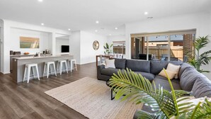 A modern living room with a grey sofa, a coffee table, and a kitchen area with a bar and stools. - City & Surf Getaway - Luxe, Spacious, Sleeps 12 (Armstrong Creek)