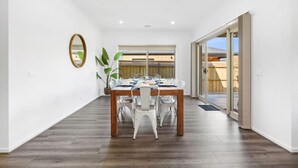 A dining area with a wooden table and white chairs, a potted plant, and a sliding glass door leading outside. - City & Surf Getaway - Luxe, Spacious, Sleeps 12 (Armstrong Creek)