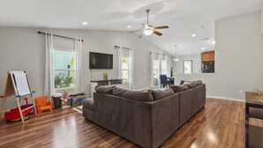 A modern living room with a sectional sofa, wooden flooring, a ceiling fan, and a flat-screen TV mounted on the wall. - Peaceful Midland Cottage- Family & Pet Friendly, Near Golf, Parks, Nature Trail & Shopping! +15% Off (Midland)