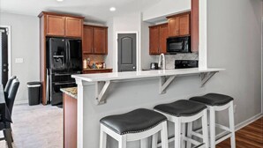 A modern kitchen with wooden cabinets, a black refrigerator, a microwave, and a white countertop. There are three white bar stools with black seats. - Peaceful Midland Cottage- Family & Pet Friendly, Near Golf, Parks, Nature Trail & Shopping! +15% Off (Midland)