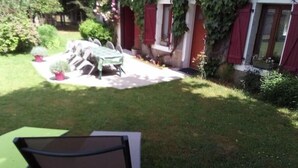 A garden with a table and chairs, a potted plant, and a red building with shutters. - Gîte "des coussieres" between peace and nature (Courteix)