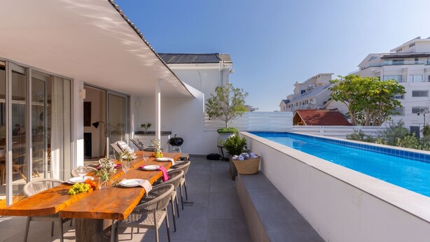 Ein moderner Essbereich im Freien mit Blick auf den Pool, ausgestattet mit einem Holztisch, der zum Essen gedeckt ist, einem Grill und einer kleinen Gartenanlage.