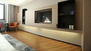 Ein modernes Hotelzimmer mit großem Flachbildfernseher an der Wand, Holzboden, einem Bett mit gemustertem Überwurf, einem kleinen Tisch mit Lampe und einem Sessel.