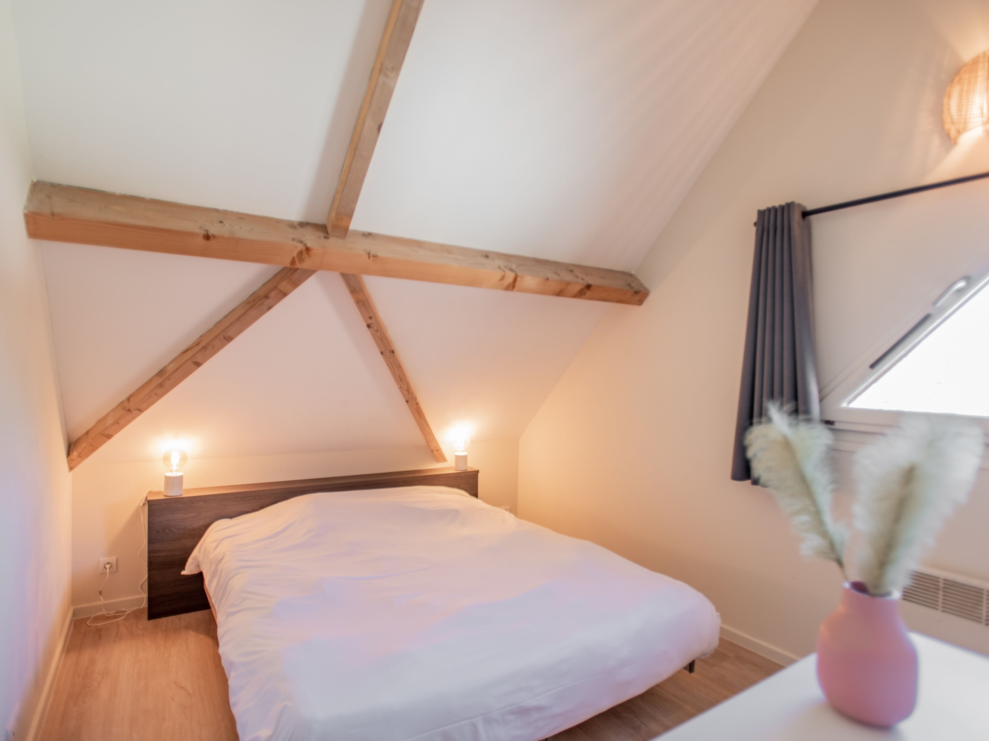 A bedroom with a wooden ceiling, a bed with white bedding, a pink vase with dried flowers, and a window with curtains.