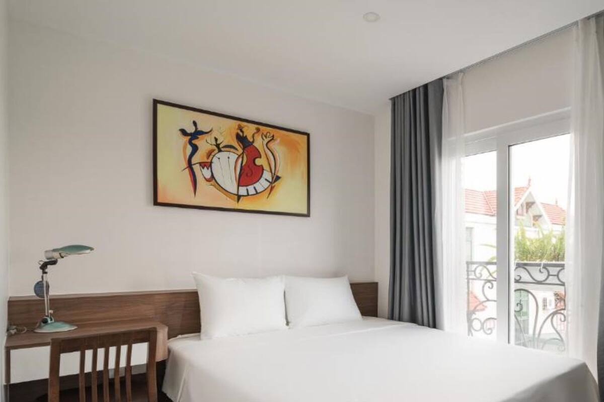 A hotel room with a bed, a desk with a lamp, a painting on the wall, and a balcony with curtains.