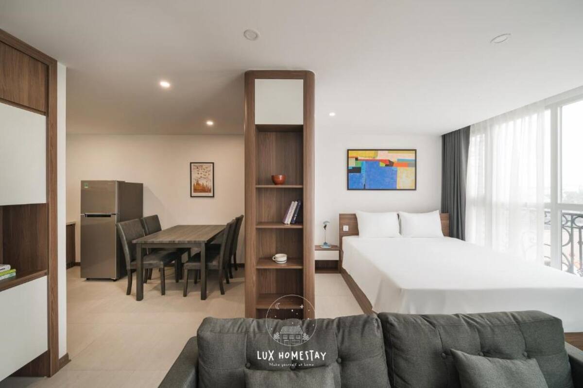 A hotel room with a bed, a dining table with chairs, a bookshelf, a refrigerator, and a large window with curtains.