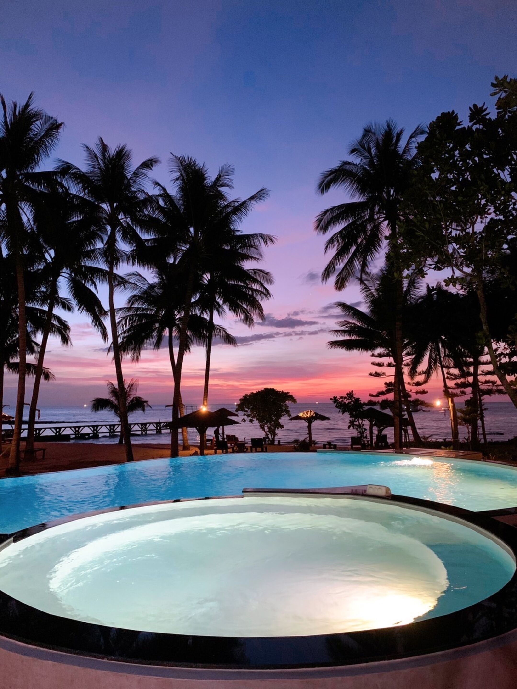 A pool area with a sunset view, surrounded by palm trees and a wooden deck.