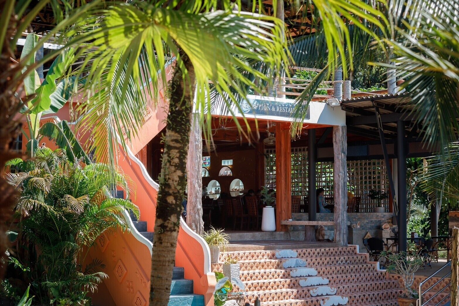 Un resort tropical con una zona de estar techada al aire libre, una escalera que lleva a una terraza de madera y exuberante vegetación.