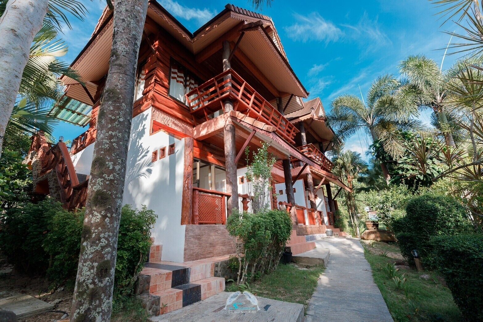 A traditional wooden house with a balcony, surrounded by lush greenery and palm trees.