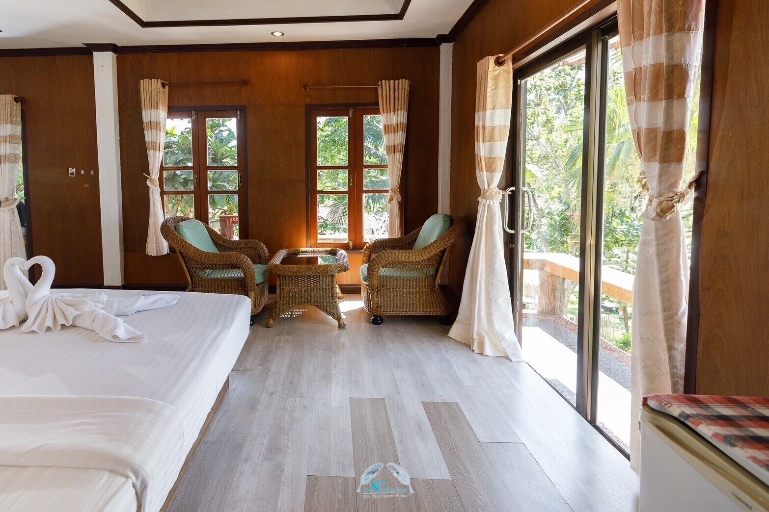 A hotel room with two beds, wicker chairs, a small table, and large windows with curtains.