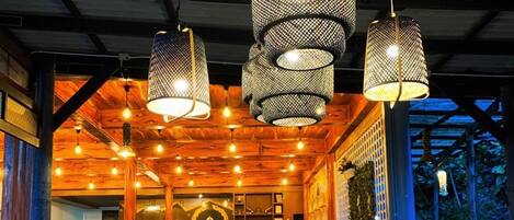 Een buitenterras met metalen stoelen en tafels, hangende netlampen en een houten plafond.