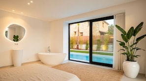 A modern bedroom with a large bed, a freestanding bathtub, a round mirror, a potted plant, and a view of a swimming pool and outdoor area.