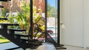 A sculpture of a figure seated on a bench by a glass door, with a view of palm trees and a building in the background.