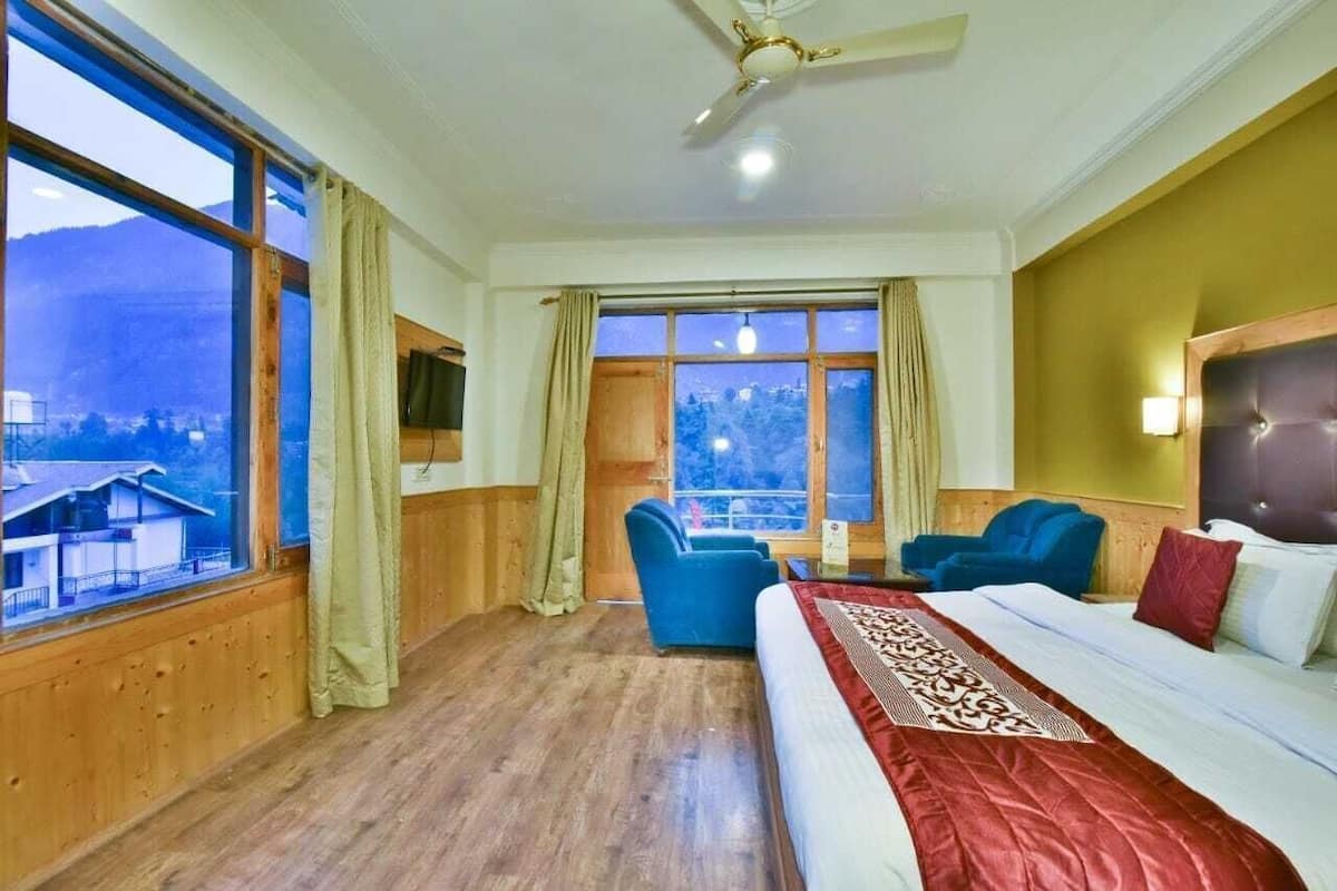 Une chambre d’hôtel avec un lit, deux chaises, une télévision et de grandes fenêtres équipées de rideaux.