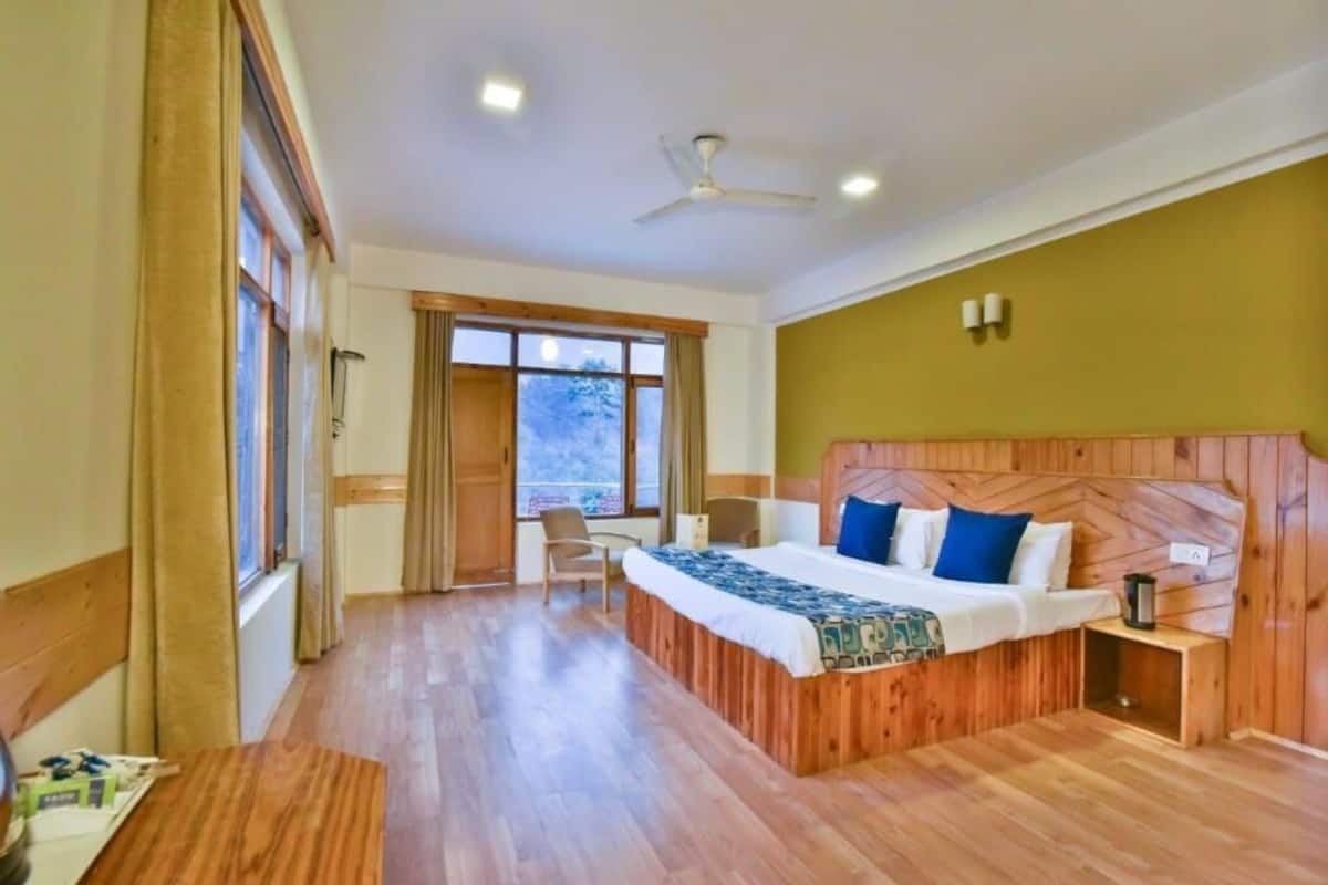 Une chambre d’hôtel avec une tête de lit en bois, un lit avec une literie bleue et blanche, un bureau avec une chaise et une fenêtre avec des rideaux.