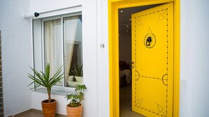 A bright yellow door with a decorative pattern, a window with white curtains, and two potted plants outside. - Peaceful corner in the center of La Marsa (Tunis)