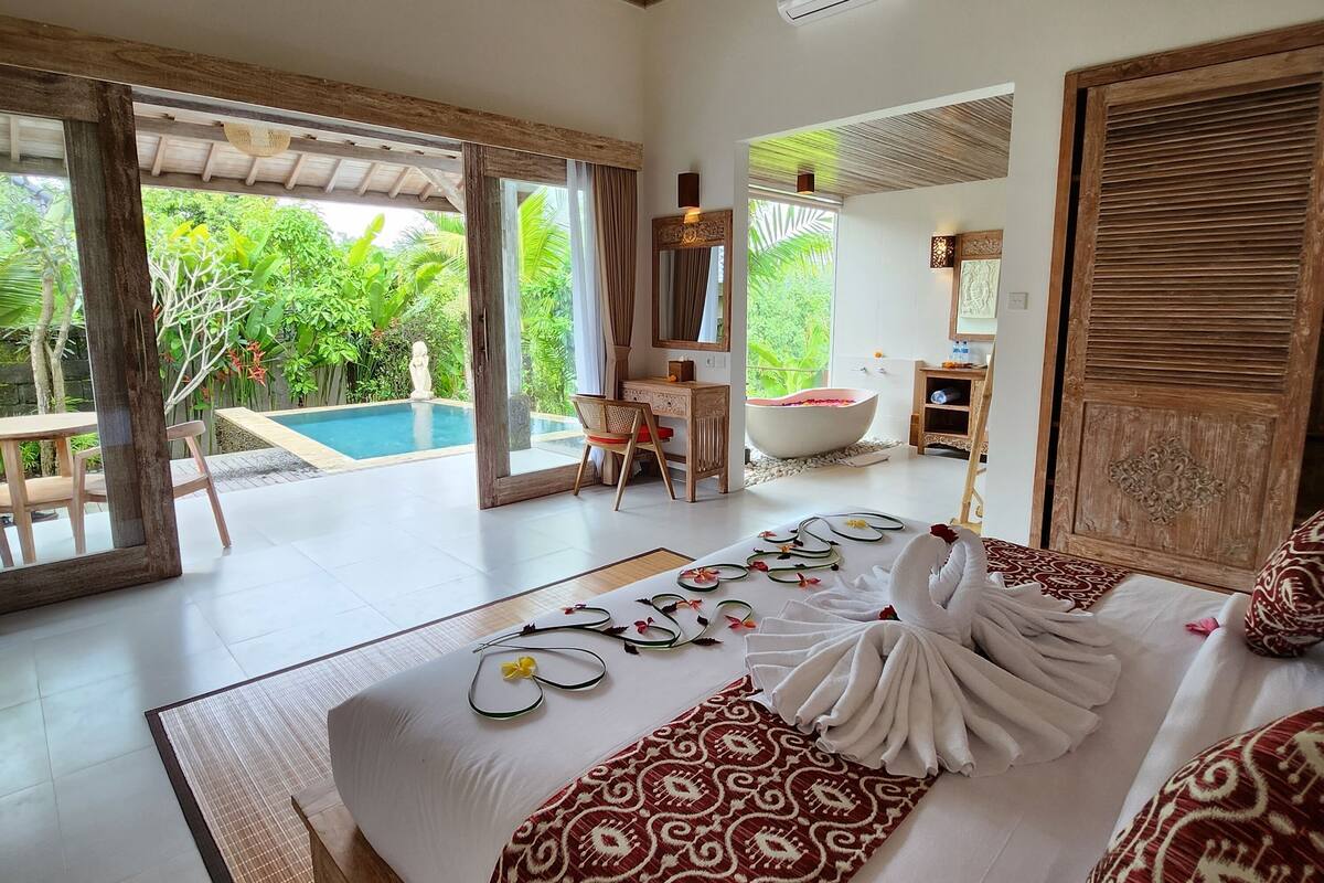 A spacious bedroom with a large bed, a sitting area with a chair, a bathtub, and a view of a swimming pool and lush greenery.
