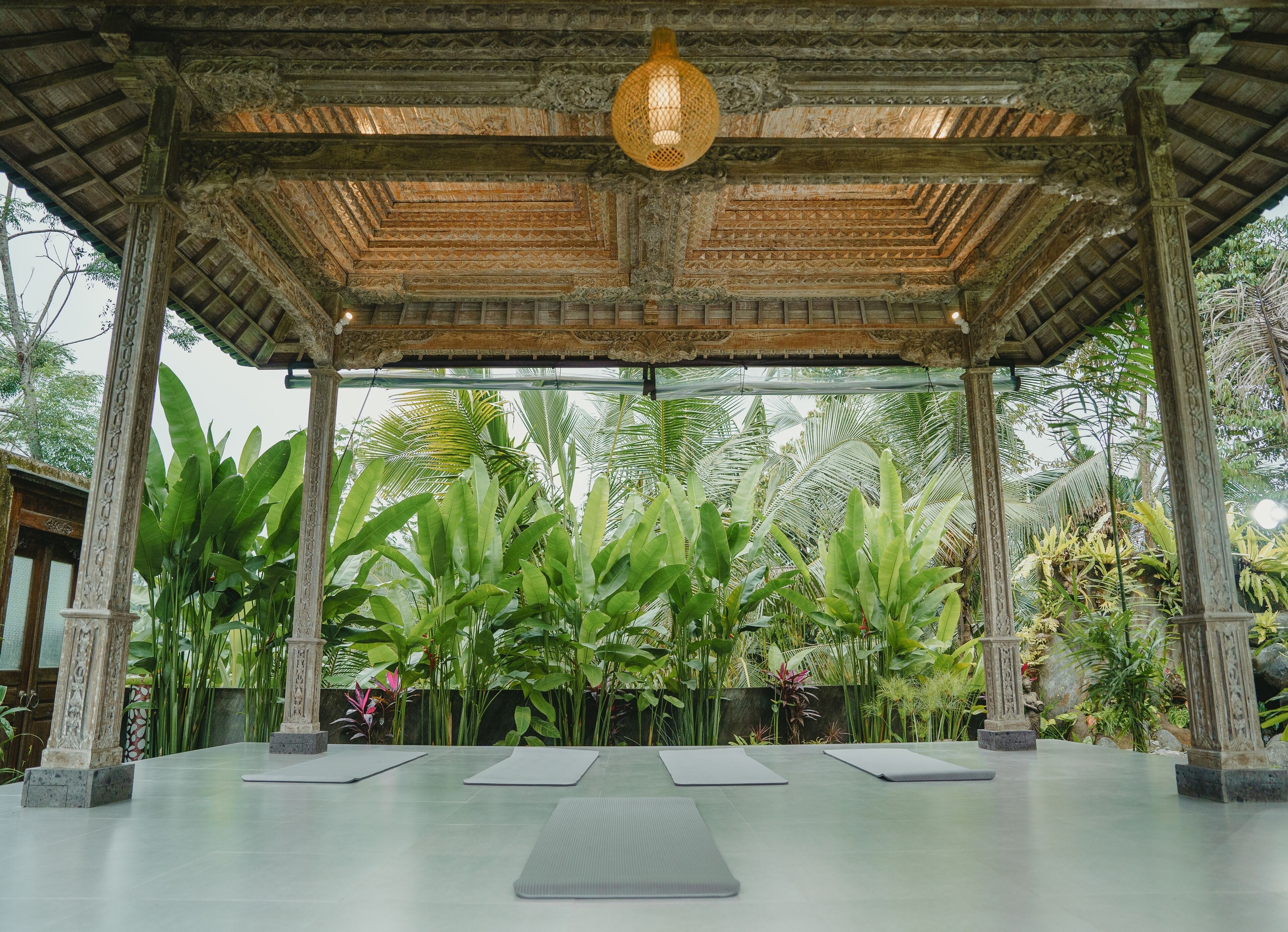 A covered outdoor area with a wooden ceiling, large green plants, and exercise mats.