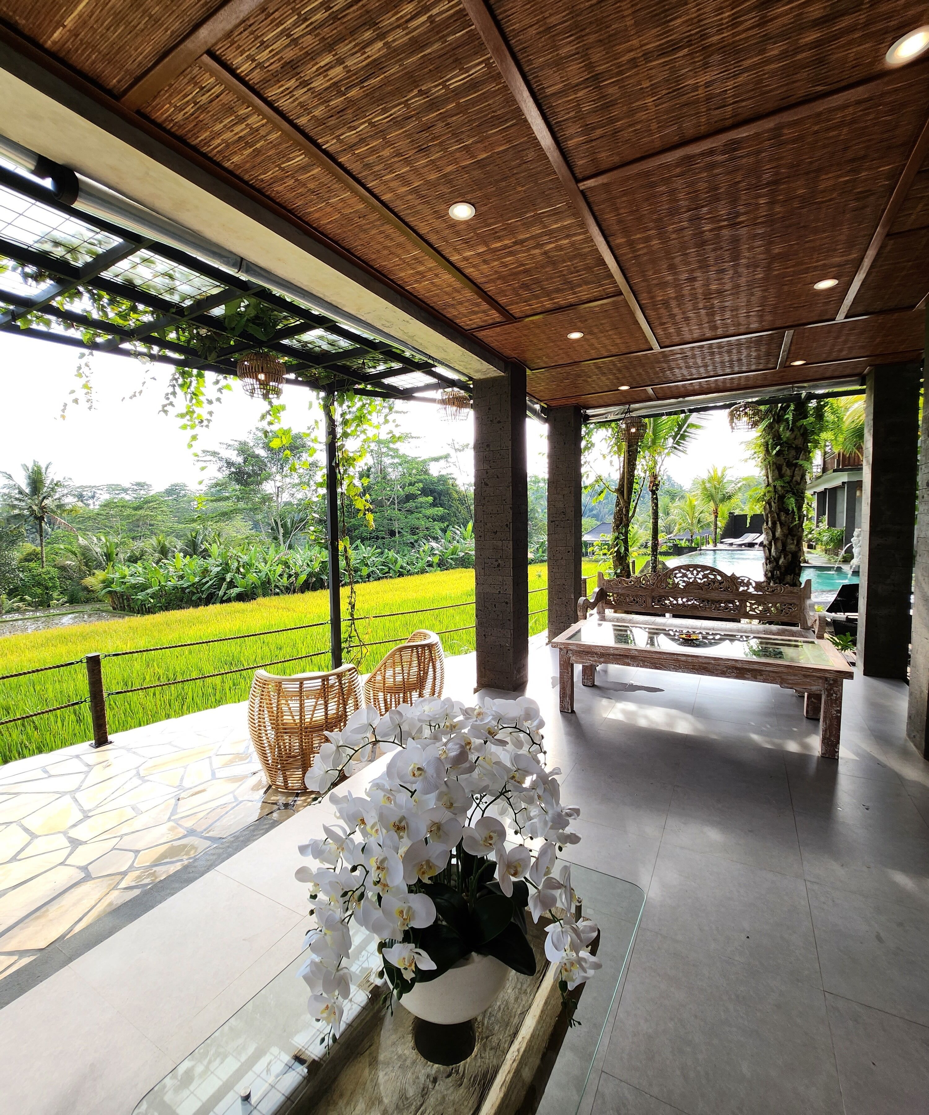 A spacious indoor area with a view of a lush green landscape and a swimming pool. There are wicker chairs and a wooden bench under a wooden ceiling.
