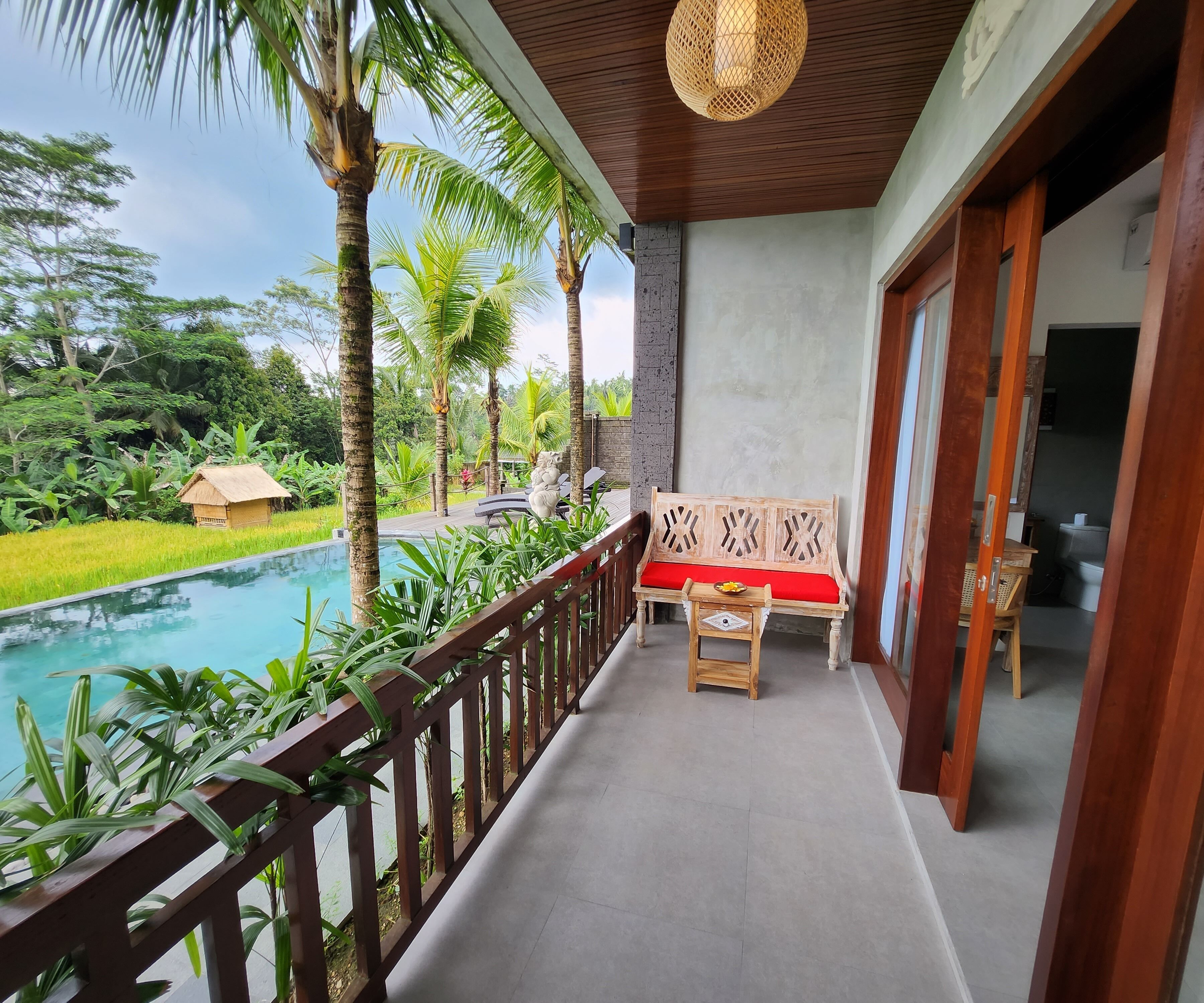 A balcony with a wooden bench and a small table, overlooking a swimming pool and lush greenery.
