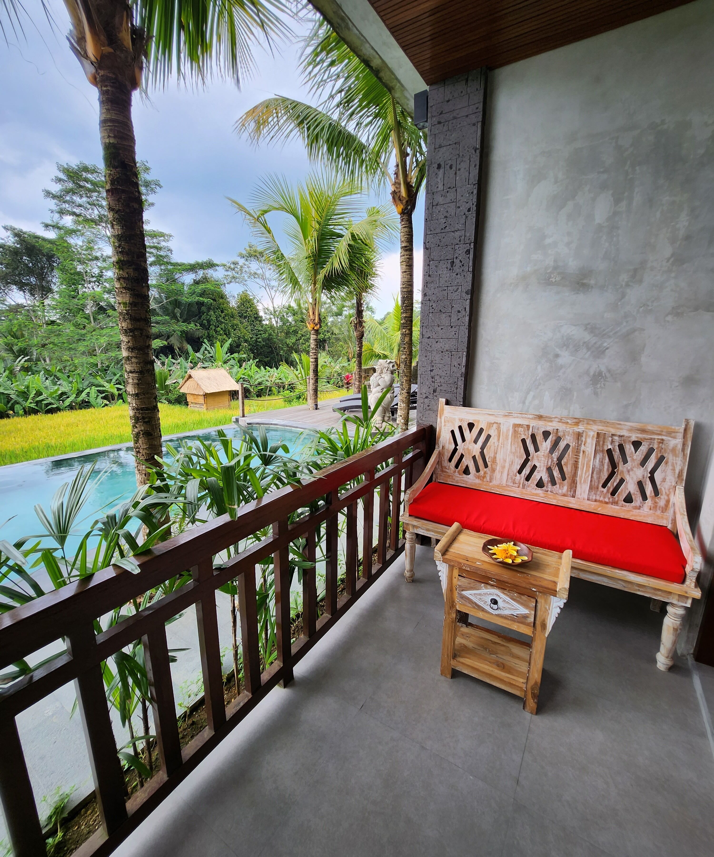 A wooden bench with a red cushion and a small table with a flower vase on a balcony overlooking a swimming pool and lush greenery.