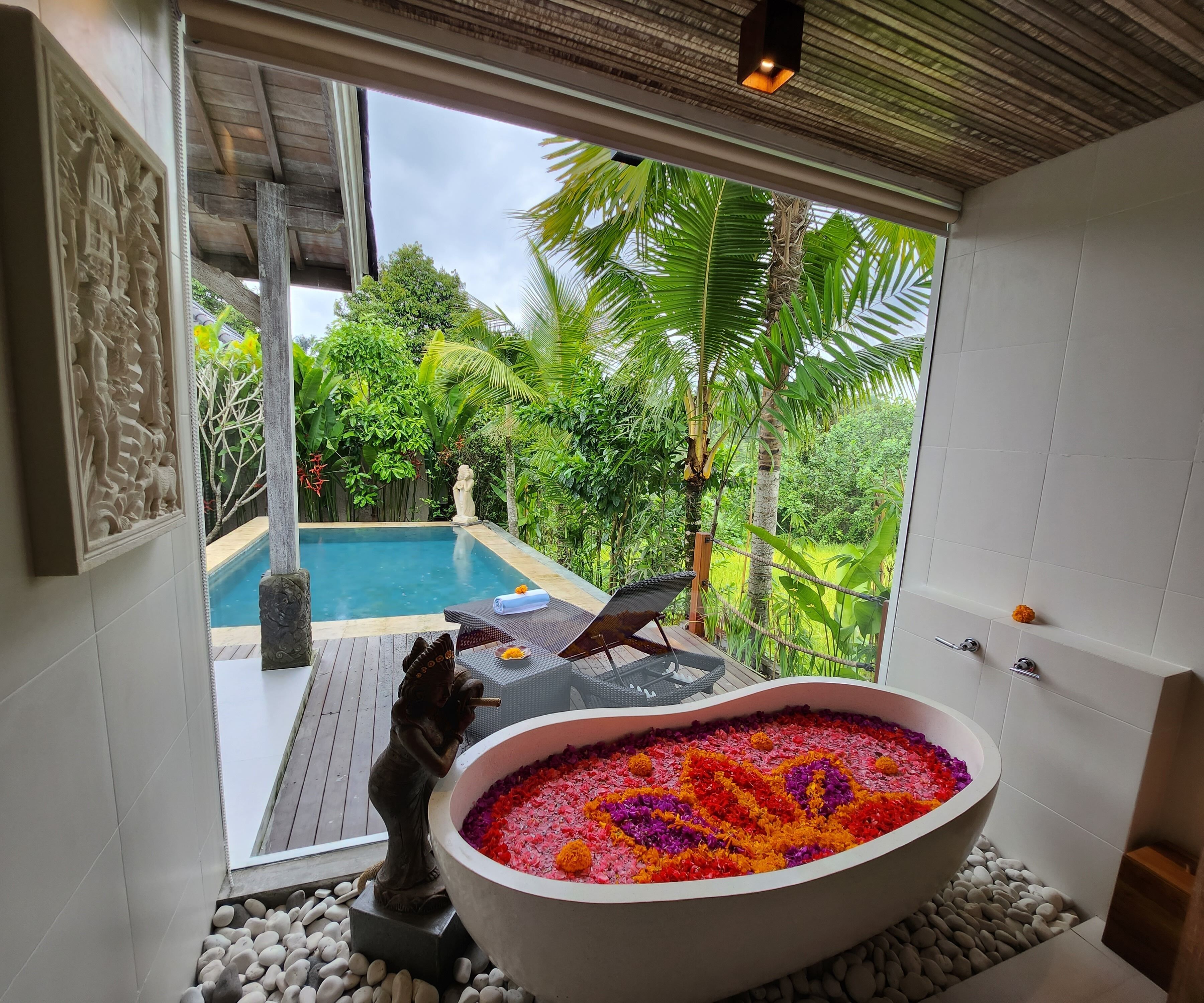 A spa area with a large bathtub filled with colorful flowers, a statue, and a view of a pool and lush greenery.