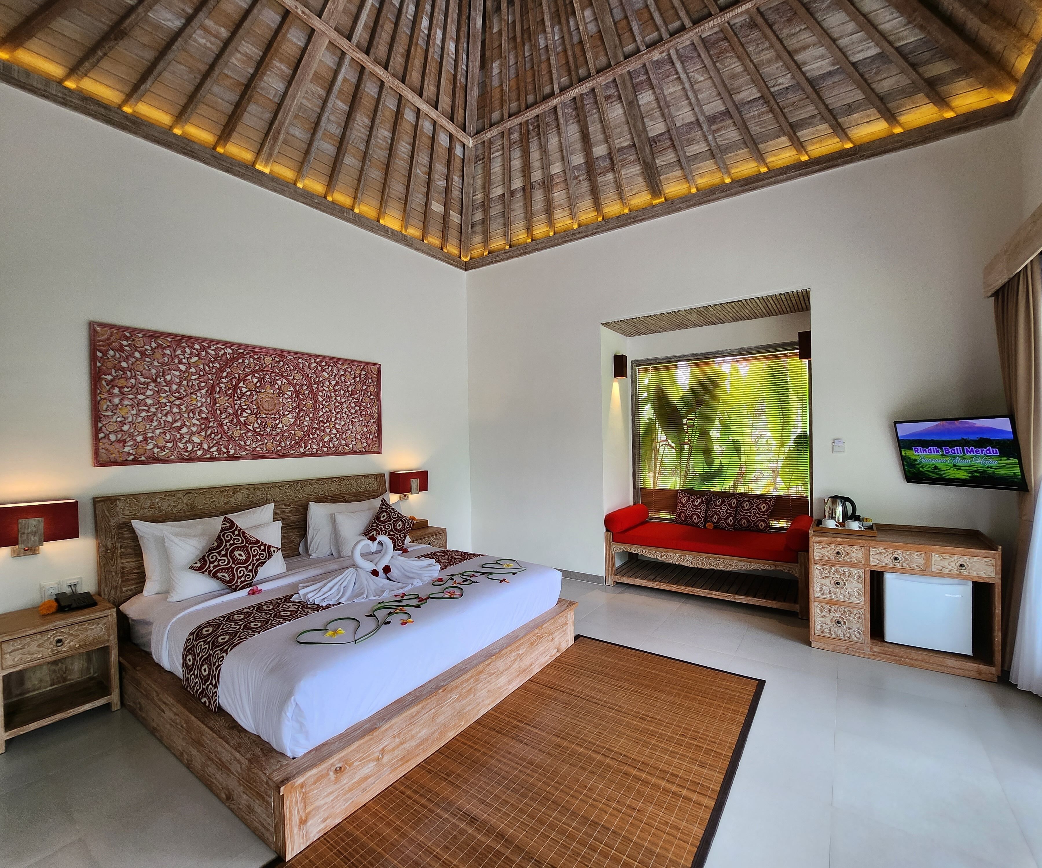 Un dormitorio con una cama grande, cabecera de madera, mesita de noche, sofá rojo, televisor y ventana con persianas.