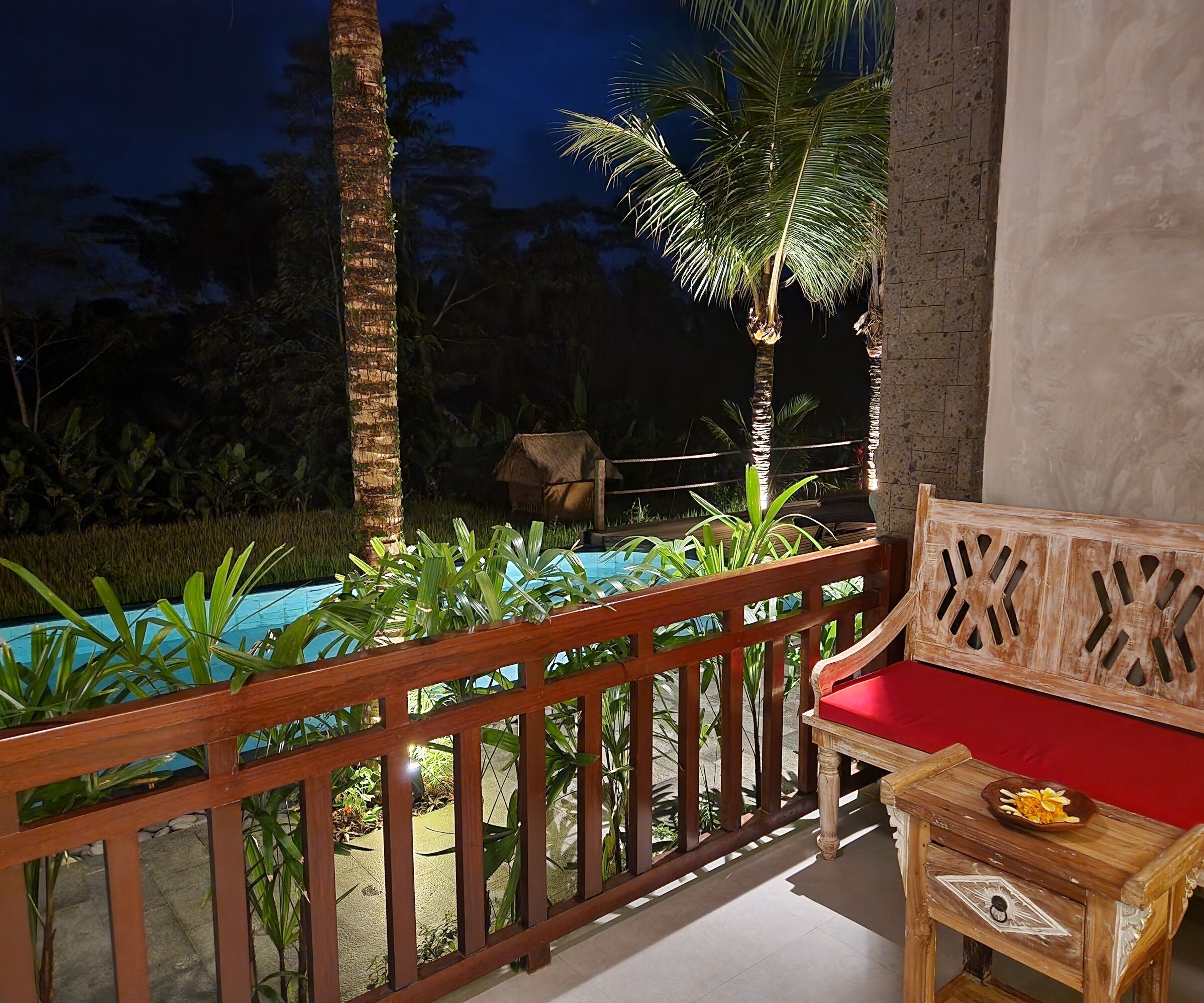 Un banco de madera con cojín rojo y una mesita con un jarrón de flores, situado en un balcón con barandilla de madera con vistas a una zona de piscina.